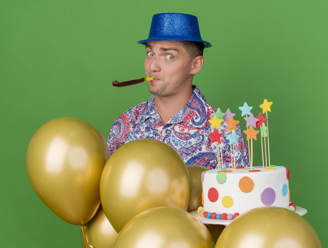 蛋糕高兴的年轻人戴着蓝色的帽子拿着吹蛋糕的吹风机站在气球后面孤立的绿色派对小伙子气球