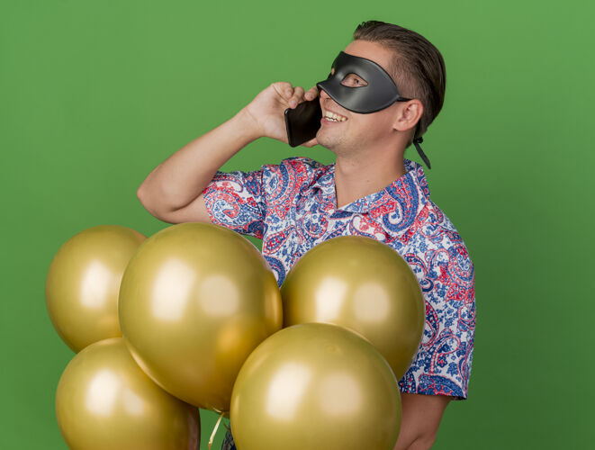 派对面带微笑的年轻人站在气球中间 戴着化装眼罩 站在一旁看派对 在绿色的隔离地带讲电话穿面具站