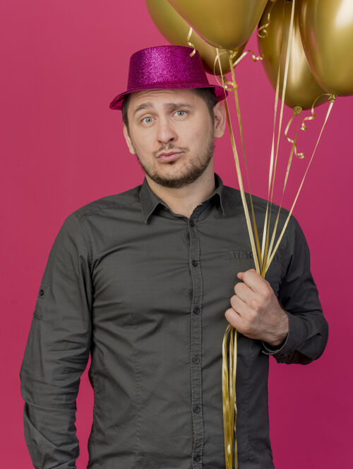 年轻人高兴的年轻人戴着粉红色的帽子拿着气球孤立在粉红色的聚会气球派对拜托