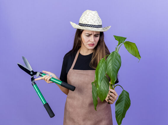 蓝色体贴美丽的园丁女孩穿着制服 戴着园艺帽 拿着剪子 看着她手上孤立的蓝色植物帽子园艺举行