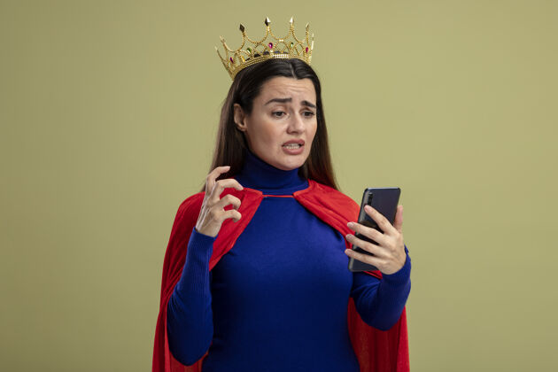 年轻关心的年轻超级英雄女孩戴着王冠 手持电话 看着橄榄绿的隔离超级英雄关心皇冠