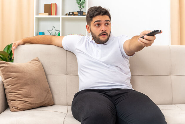 生活穿着休闲服的年轻人拿着电视遥控器看电视 兴致勃勃地在家里度过周末 坐在客厅里的沙发上周末消费观看