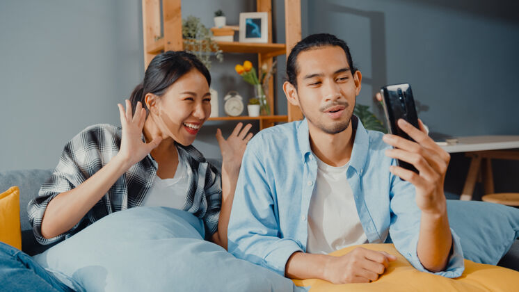 妻子快乐的亚洲年轻夫妇男女坐在沙发上使用智能手机与朋友和家人进行facetime视频通话笑享受娱乐