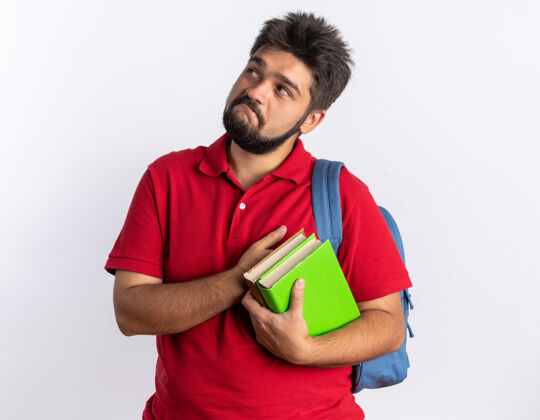 拿着年轻的留着胡子的学生 穿着红色马球衫 背着背包 举着笔记本 脸上带着怀疑的表情 站在白色的背景上仰望着胡子表情怀疑