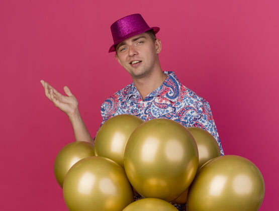 气球高兴的年轻人戴着粉红色的帽子站在气球后面 用手指着旁边孤立的粉红色壁板请站