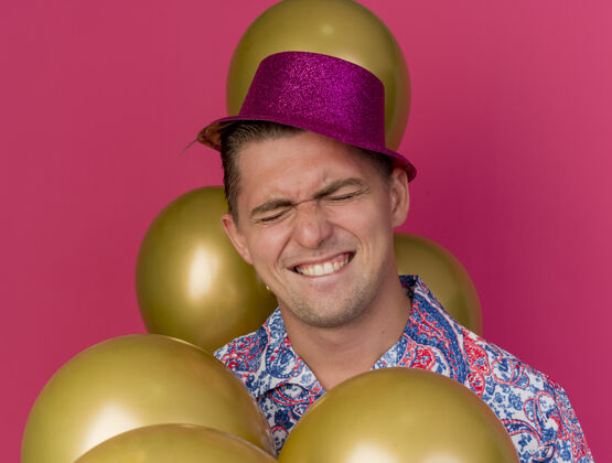 欢乐快乐的年轻人戴着粉色帽子闭着眼睛站在粉红色的气球中间年轻站男人