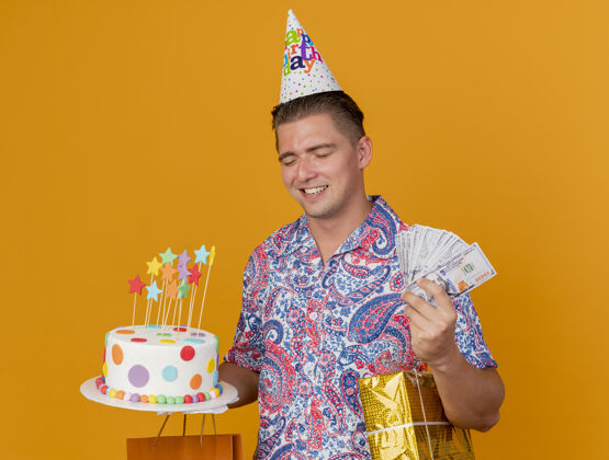 聚会笑容满面的年轻人 戴着生日帽 闭着眼睛 手里拿着蛋糕 蛋糕上的礼物和钱被隔离在橘子上家伙年轻眼睛