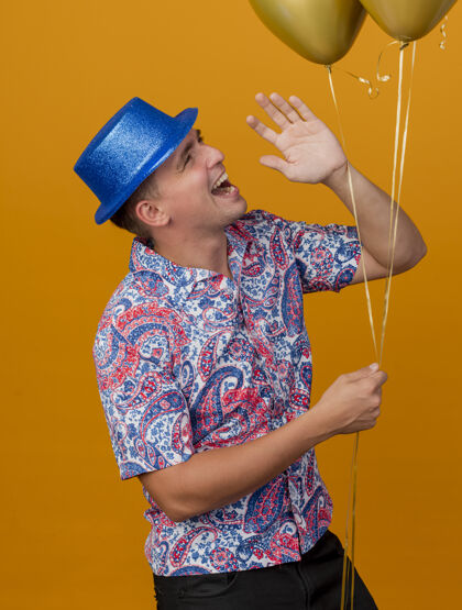 戴着带着微笑的年轻人戴着蓝色的帽子拿着气球 在橘子上叫着一个孤立的人小伙子派对有人
