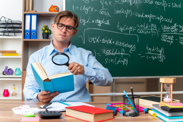 教室年轻的男老师戴着眼镜坐在书桌旁 拿着书和笔记 透过放大镜看着教室黑板前严肃的书本老师眼镜书桌