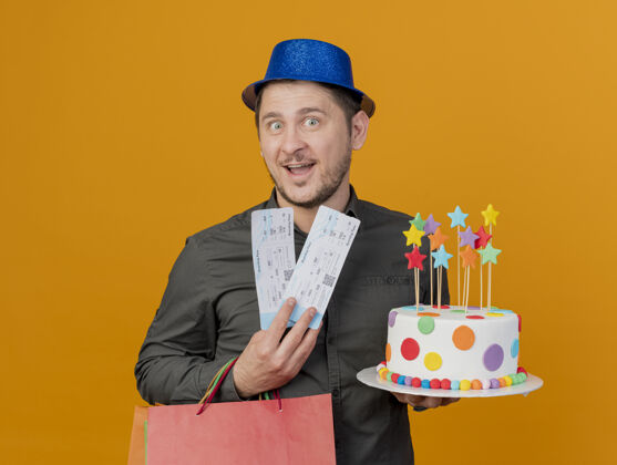 蓝色带着微笑的年轻人戴着蓝色的帽子拿着蛋糕和礼包 票子被隔离在橙色上包穿礼物