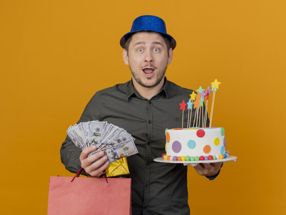 年轻人惊讶的年轻人戴着蓝色帽子拿着蛋糕和礼品袋 现金孤立在橙色穿上现金派对