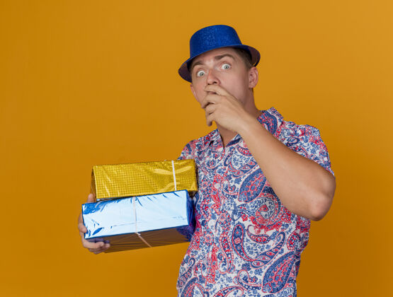 橙色惊讶的年轻人戴着蓝色的帽子拿着礼物盒抓住下巴孤立在橙色帽子男人盒子