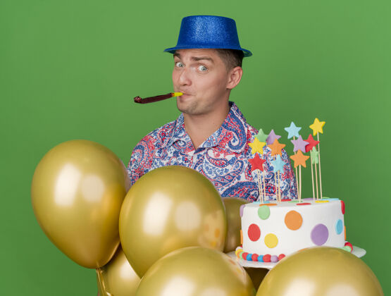 蛋糕高兴的年轻人戴着蓝色的帽子拿着吹蛋糕的吹风机站在气球后面孤立的绿色派对小伙子气球