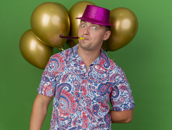 派对惊讶的年轻人戴着粉红色的帽子站在气球前 吹着绿色的派对吹风机气球年轻吹