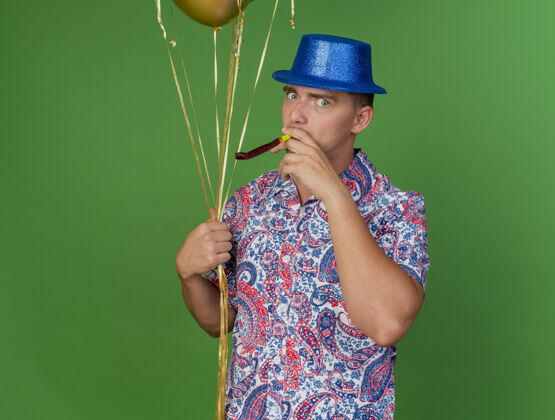 蓝色高兴的年轻人戴着蓝帽子 拿着气球吹着聚会的吹风机孤立在绿色绿色气球戴