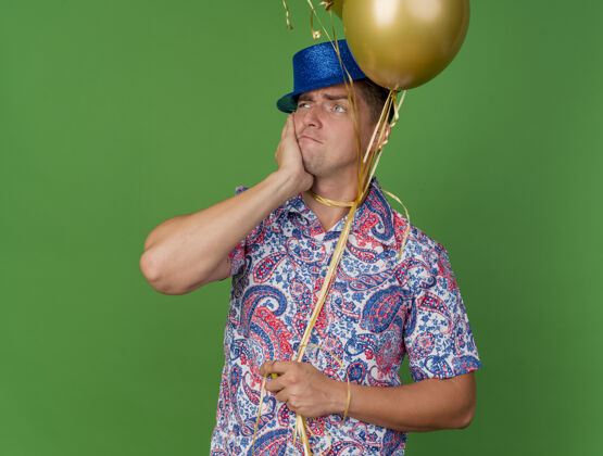 蓝色一个体贴的年轻人 戴着蓝色帽子 手里拿着气球 脖子上系着气球 手放在脸上 脸上贴着绿色年轻领带脸颊