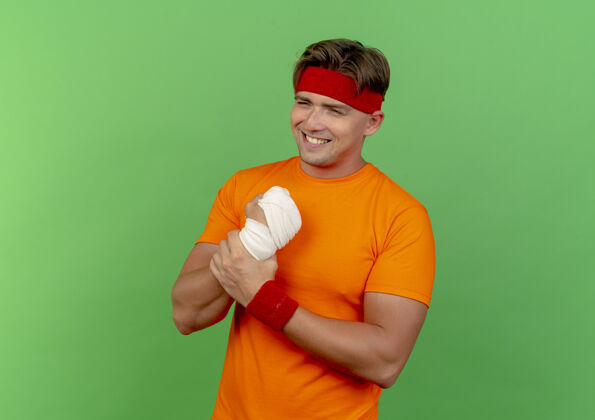 抱着面带微笑的年轻英俊的运动型男子戴着头带和腕带 用绷带包裹着他受伤的手腕 隔离在绿色的墙上市民绿色腕带