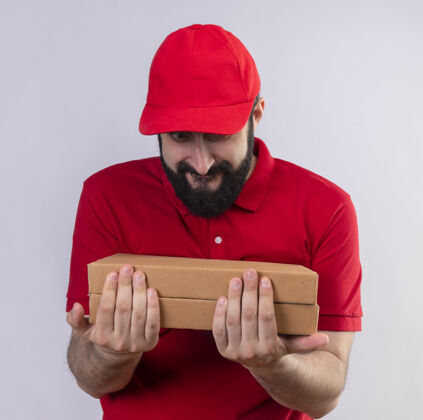人年轻帅气的送货员穿着红色制服 戴着帽子 看着隔离在白墙上的披萨盒姿势感觉脸
