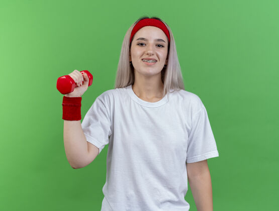 腕带微笑的年轻运动女性戴着背带 戴着头带和腕带 把哑铃孤立地放在绿色的墙上女孩哑铃头带
