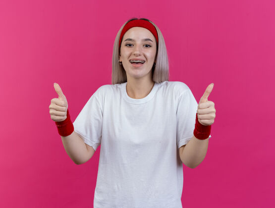 运动兴奋的年轻运动女性戴着牙套戴着头带和腕带两只手竖起大拇指孤立在粉红色的墙上女人人拇指