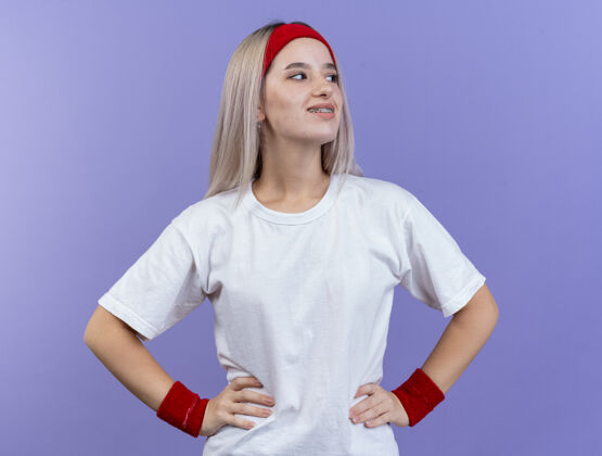 腕带带着背带 戴着头带和腕带 面带微笑的年轻运动女性把手放在腰上 看着紫色墙上孤立的一面穿人腰