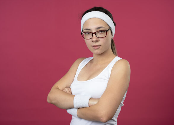 光学自信的年轻运动女性戴着眼镜戴着头带和腕带交叉双臂站在粉红色的墙上腕带手臂女人