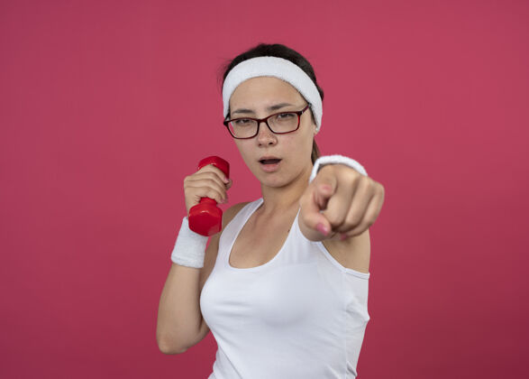 举行自信的年轻运动女性戴着眼镜 戴着头带和腕带 举着哑铃 指着隔离在粉色墙上的前面运动女人人