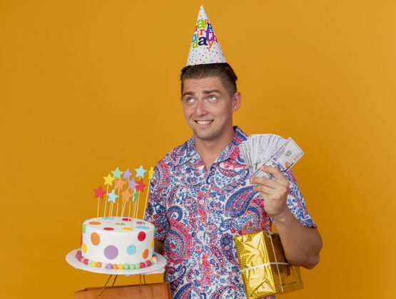 橘子关心派对的年轻人戴着生日帽 手里拿着蛋糕 手里拿着礼物和现金 孤立地站在橘子上派对现金年轻人