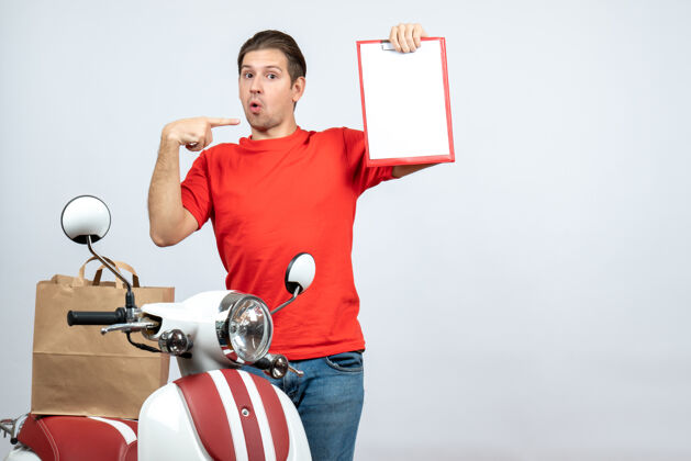 成人前视图混乱的送货员穿着红色制服站在滑板车附近 显示文件指向自己的白色背景微笑健身送货员