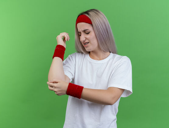 女孩带着背带 戴着头带和腕带 疼痛的年轻运动女性把手放在隔离在绿墙上的手肘上头带肘部年轻