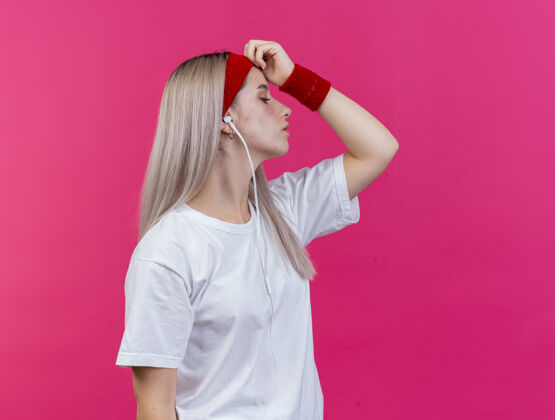 女孩自信的年轻运动女性戴着耳机戴着牙套戴着头带和腕带站在一边把手放在隔离在粉色墙上的前额上头带佩戴手