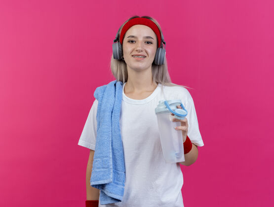 女孩微笑的年轻运动女性戴着耳机戴着牙套戴着头带和腕带肩上拿着水瓶和毛巾隔离在粉红色的墙上运动持有背带