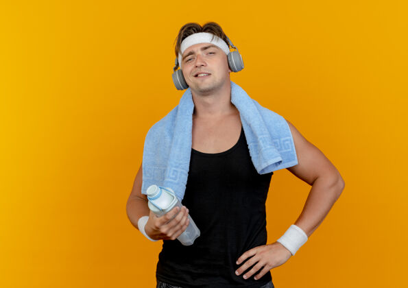 围着高兴的年轻英俊的运动型男子戴着头带和腕带和耳机 脖子上围着毛巾拿着水瓶 手放在腰上 隔离在橙色的墙上脸瓶子姿势