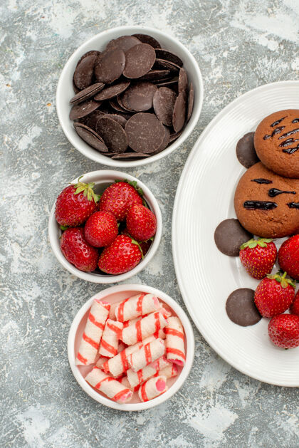 水果上半部分是白色椭圆形盘子上的巧克力饼干草莓和圆形巧克力 灰白色地面上的碗里有糖果草莓巧克力浆果健康新鲜