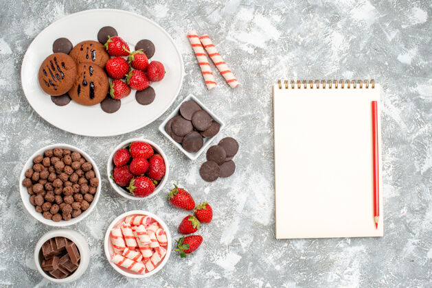 食物俯视饼干草莓和圆形巧克力放在椭圆形的盘子里 碗里放着糖果草莓巧克力麦片 灰白色的桌子上放着一个带铅笔的笔记本甜点糖果碗