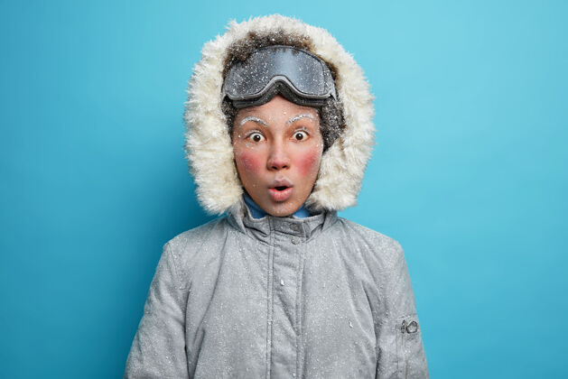 夹克穿着灰色夹克 戴着风帽和滑雪护目镜 脸冻得通红的女滑雪者惊呆了美丽服装冬天
