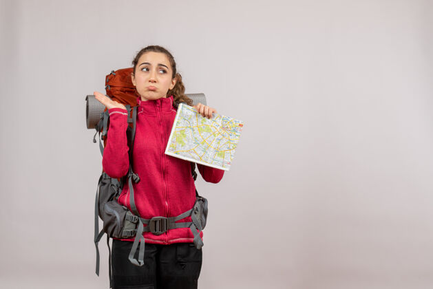 快乐困惑的年轻旅行者的正面图 背着大背包 在灰色的墙上举着地图可爱前面地图