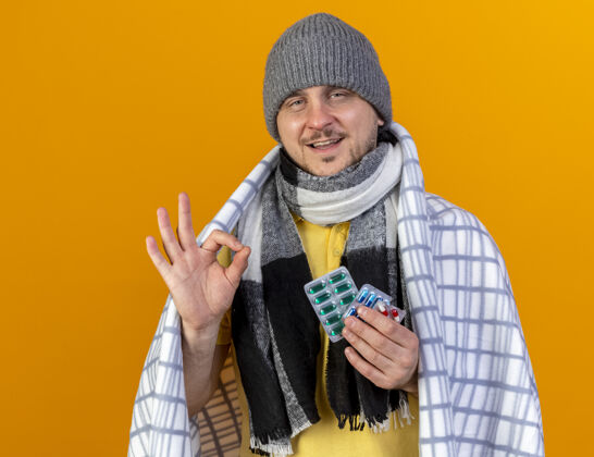 男人高兴的年轻金发病斯拉夫男子戴着冬天的帽子和围巾手势好的手势 并持有一包医药药片包裹在格纹隔离在橙色墙上的复制空间请包包裹