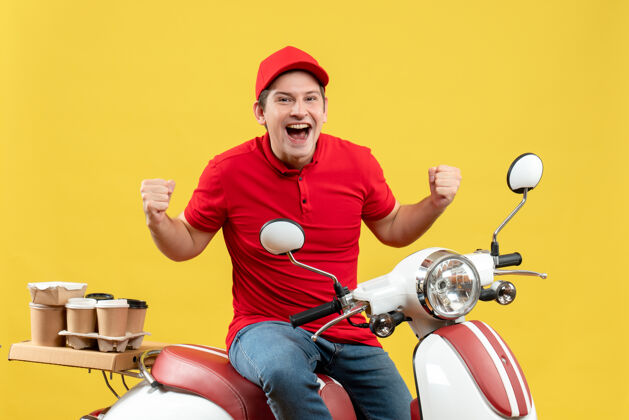 人俯视图：微笑的年轻人穿着红色上衣 戴着帽子 坐在踏板车上传递着命令 在黄色的墙上感到快乐年轻人感情快乐