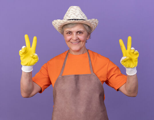 园艺微笑的上了年纪的女园丁戴着园艺帽和手套 在紫色上打着胜利的手势女胜利帽子