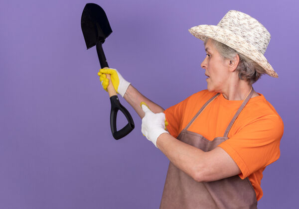 帽子自信的上了年纪的女园丁戴着园艺帽和手套拿着铲子指着紫色的一边看点黑桃手套