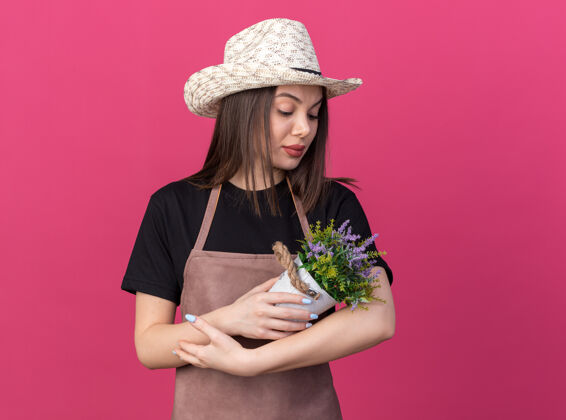 园艺自信漂亮的白人女园丁戴着园艺帽捧着粉色的花盆看着自信帽子花园