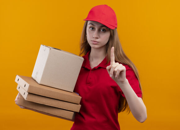 提高一个穿着红色制服 表情严肃的年轻送货女孩 手拿着盒子和包裹 手指高举在孤立的橙色空间里女孩红色盒子