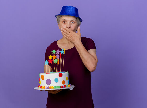 穿惊讶的戴着派对帽的老太太把手放在嘴上 拿着生日蛋糕孤立地放在紫色的墙上 留着复制空间生日帽子举行