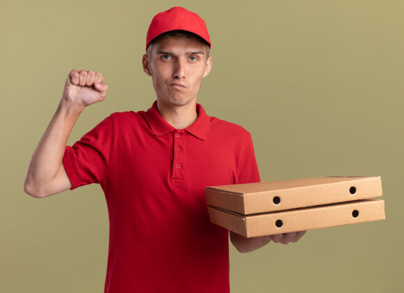拳头自信的年轻金发送货员举起拳头 拿着橄榄绿的比萨饼盒金发披萨向上