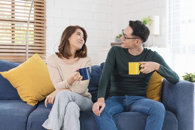 年轻快乐的亚洲夫妇在家里一起在沙发上度过周末 放松和享受喝咖啡在一起沙发房子