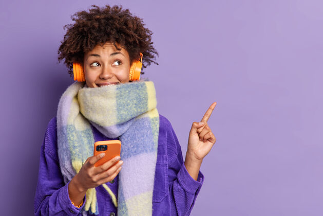 美国快乐体贴的美国黑人妇女裹着围巾拿着手机上网通过立体声耳机听音乐右上角的点显示文本的空白模特空乐趣