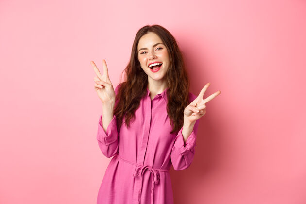 快乐积极漂亮的女孩穿着时尚的连衣裙 展现v字的和平和微笑的幸福 在粉红色的墙壁附近摆姿势和平魅力积极