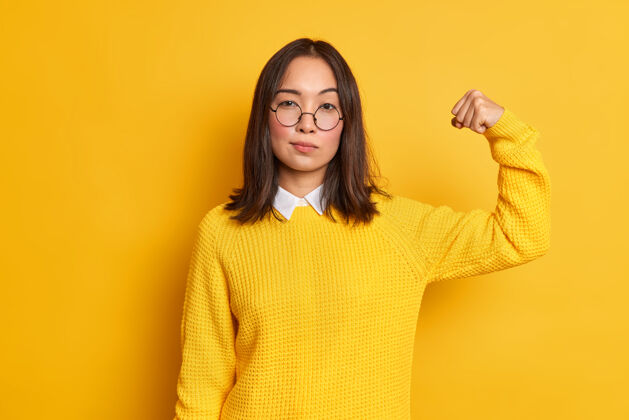 女性严肃的黑发亚洲女人的照片举起手臂 显示她的力量有强壮的肌肉站立自信的室内穿着黄色毛衣和圆眼镜女人的力量概念人呼吁二头肌
