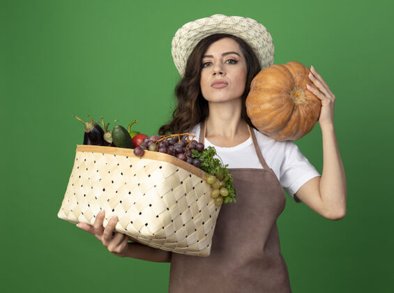 南瓜自信的年轻女园丁穿着制服 戴着园艺帽 肩上扛着菜篮和南瓜 隔离在绿色的墙上 留着复制空间肩膀蔬菜拿着
